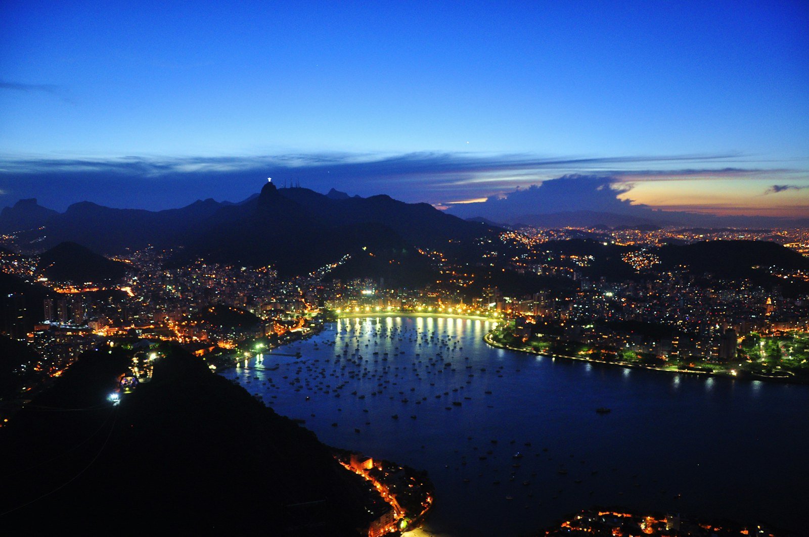 Vista noturna do Rio de Janeiro com bairros e vias iluminados ao redor da Baía de Guanabara.